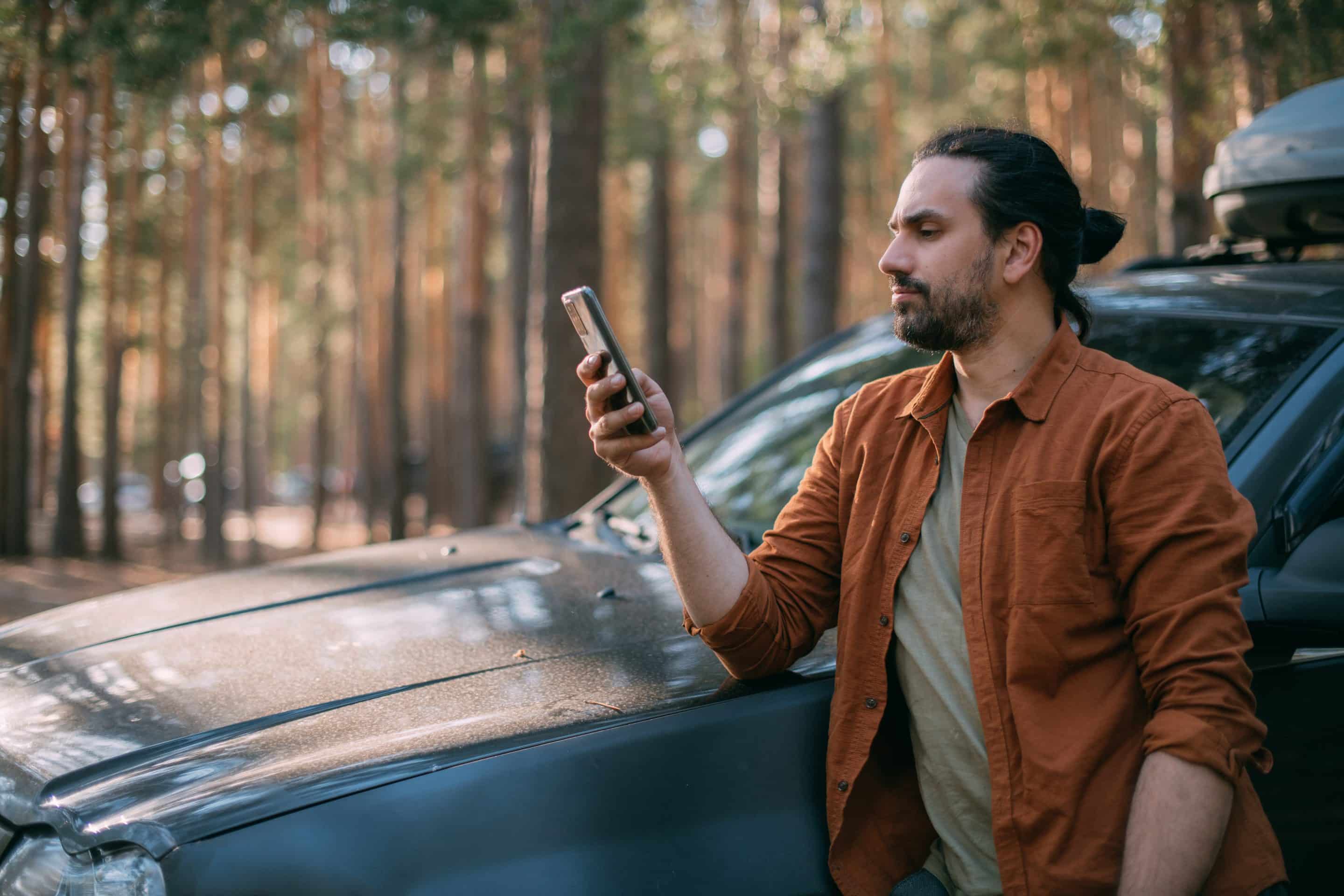 Un homme appuyé sur le capot d'une voiture en train de parler au téléphone dans une forêt