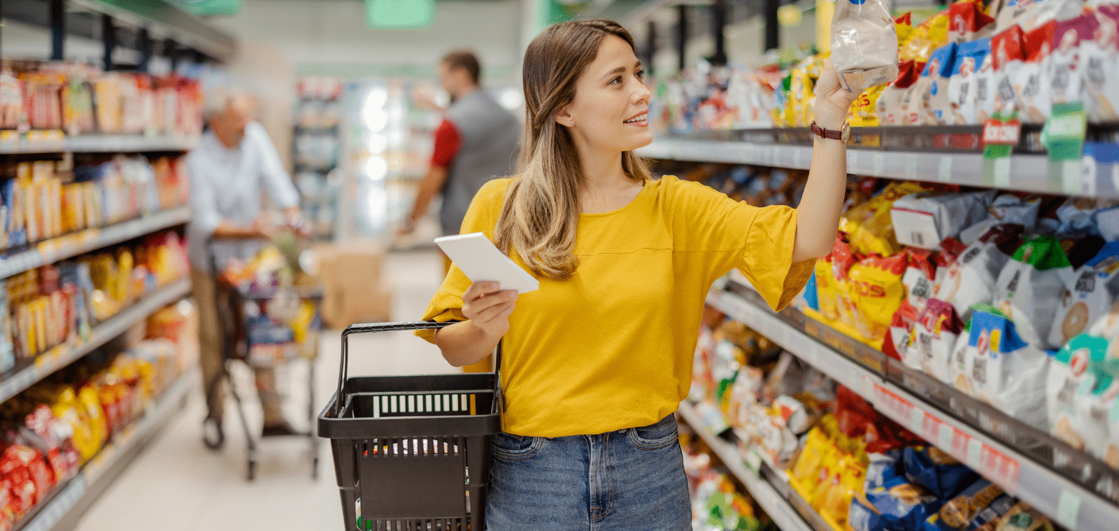 Une femme en chemise jaune fait ses courses dans un supermarché, tenant une liste dans une main et un produit dans l'autre, tandis que d'autres clients font leurs achats en arrière-plan.